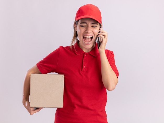 Jovem bonita entregadora de uniforme irritada segurando uma caixa de papelão e gritando com alguém no telefone, isolado na parede branca