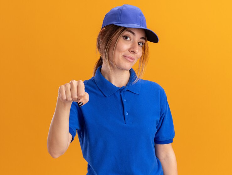 Foto grátis jovem bonita entregadora de uniforme satisfeita com o punho cerrado e olhando para a frente, isolada na parede laranja
