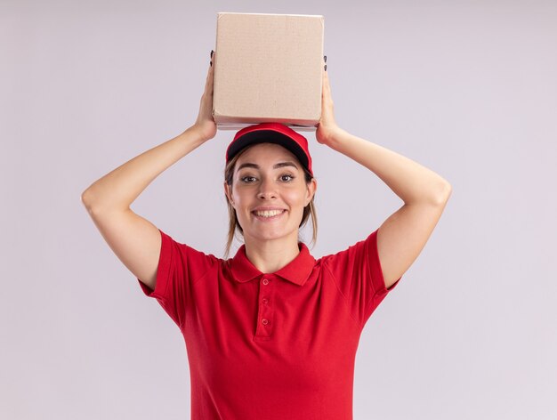 Jovem bonita entregadora de uniforme sorridente segurando uma caixa de papelão isolada na parede branca
