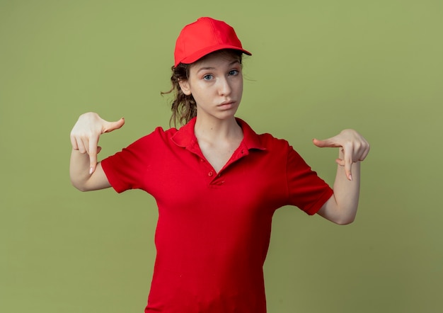 Jovem, bonita, entregadora, impressionada, com uniforme vermelho e boné apontando para baixo, isolado em um fundo verde oliva com espaço de cópia