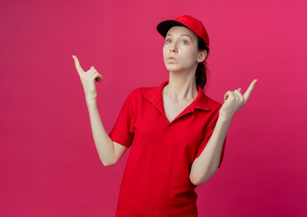 Jovem, bonita, entregadora, impressionada, com uniforme vermelho e boné, olhando em linha reta e apontando para cima, isolada em um fundo carmesim com espaço de cópia