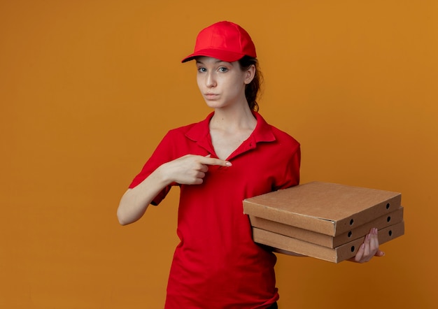 Jovem, bonita, entregadora, impressionada, com uniforme vermelho e boné, segurando e apontando para pacotes de pizza isolados em um fundo laranja com espaço de cópia