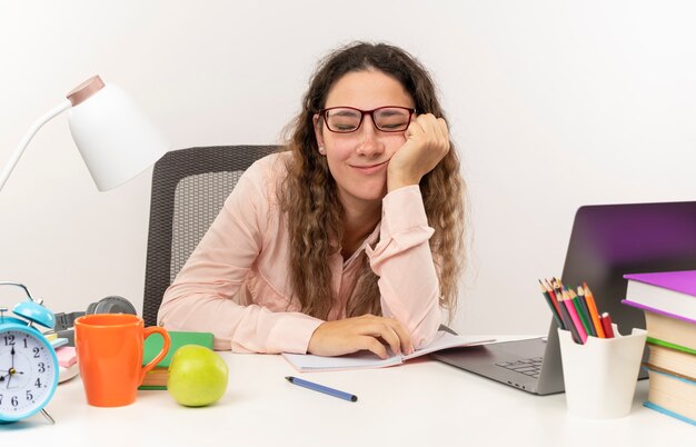 Jovem bonita feliz usando óculos, sentada na mesa com as ferramentas da escola, fazendo sua lição de casa, colocando a mão na bochecha com os olhos fechados, isolados na parede branca