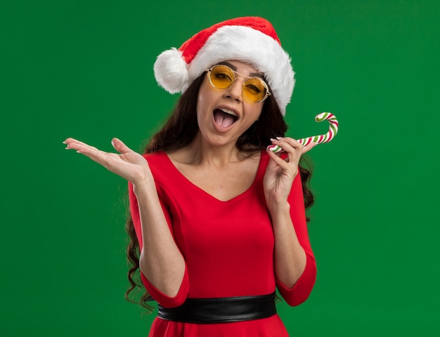 Foto grátis jovem bonita impressionada com chapéu de papai noel e óculos segurando um pirulito de natal, olhando para a câmera, mostrando a mão vazia isolada no fundo verde