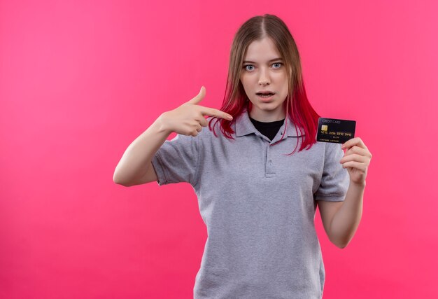 Jovem bonita impressionada com uma camiseta cinza apontando para um cartão de crédito na mão na parede rosa isolada