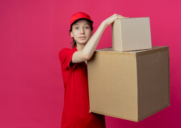 Jovem bonita impressionada com uma entregadora de uniforme vermelho e boné segurando caixas de papelão, olhando para a câmera isolada em um fundo carmesim com espaço de cópia