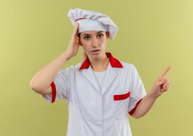 Jovem bonita impressionada com uniforme de chef, colocando a mão na cabeça e apontando para o lado isolado na parede verde