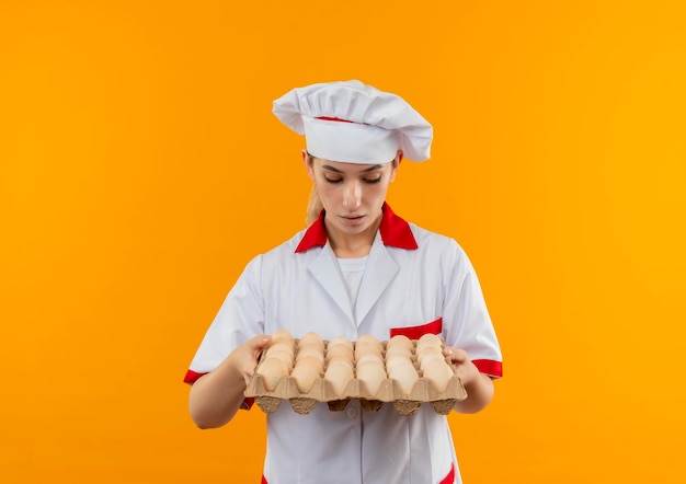 Foto grátis jovem bonita impressionada com uniforme de chef segurando e olhando para uma caixa de ovos isolada na parede laranja