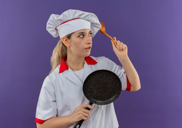 Jovem bonita impressionada com uniforme de chef segurando uma frigideira e uma colher, olhando para o lado isolado na parede roxa