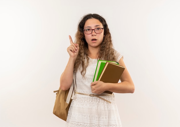 Jovem bonita impressionada de colegial de óculos e bolsa com as costas segurando livros levantando o dedo isolado no branco