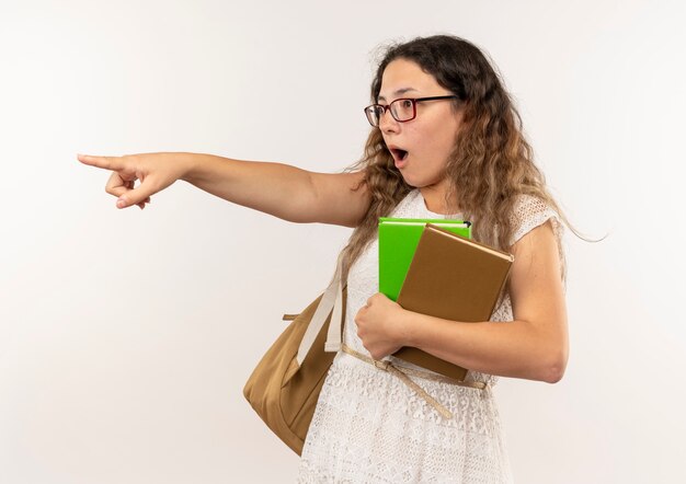 Jovem bonita impressionada de colegial de óculos e bolsa traseira segurando livros olhando e apontando para o lado isolado no branco