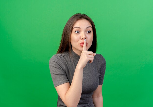 Jovem bonita impressionada olhando em linha reta e gesticulando para o silêncio isolado em um fundo verde com espaço de cópia