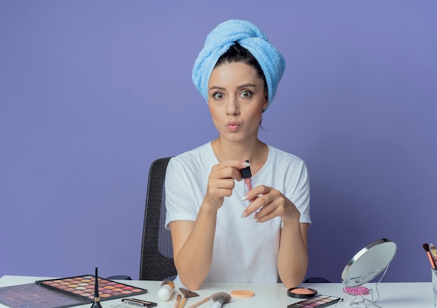 Jovem bonita impressionada sentada à mesa de maquiagem com ferramentas de maquiagem e com uma toalha de banho na cabeça segurando gloss