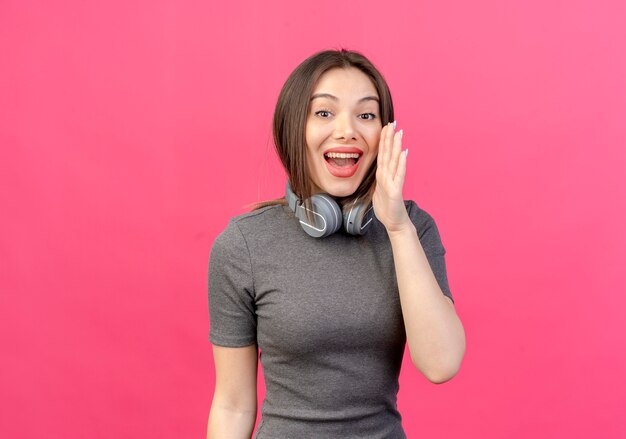 jovem bonita impressionada usando fones de ouvido no pescoço e sussurrando na frente