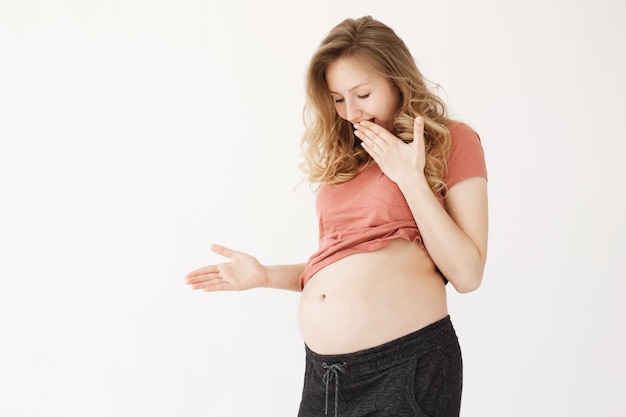 Jovem bonita loira grávida mulher bonita com cabelo comprido em roupas malucas, fechando a boca com a mão, olhando para a barriga quando o bebê empurra o estômago com a perna.