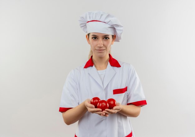 Jovem bonita satisfeita com o uniforme de chef segurando tomates e parecendo isolados no espaço em branco