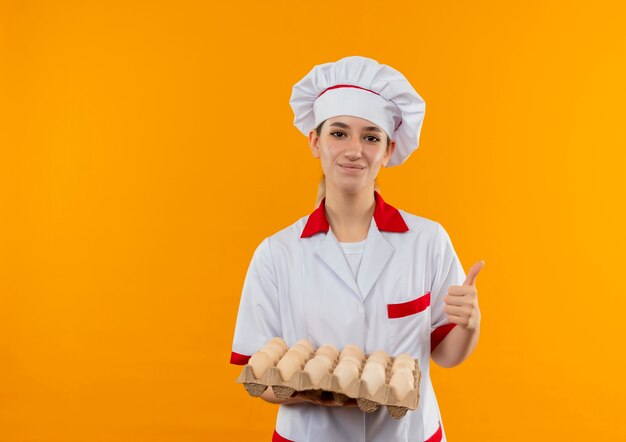 Jovem bonita satisfeita com o uniforme do chef segurando uma caixa de ovos e mostrando o polegar isolado no espaço laranja