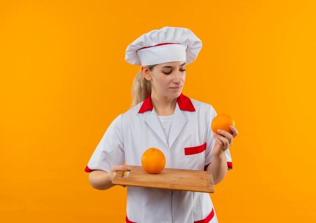 Jovem bonita satisfeita com o uniforme do chef segurando uma laranja e uma tábua olhando para a laranja isolada no espaço laranja