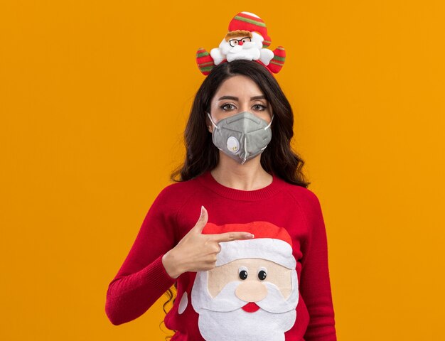 Jovem bonita séria com bandana de Papai Noel e suéter com máscara protetora apontando para o lado isolado na parede laranja