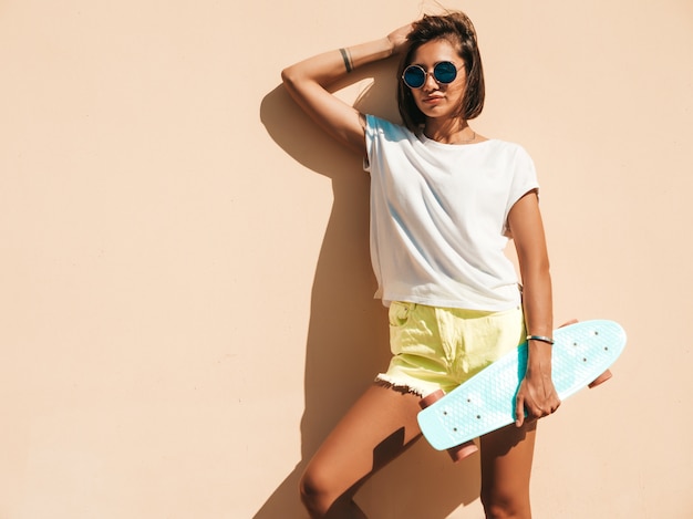 Jovem bonita sexy sorridente mulher hipster em óculos de sol. Garota da moda no verão t-shirt e shorts. Mulher positiva com centavo azul skate posando na rua perto da parede