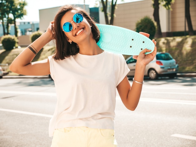 Jovem bonita sexy sorridente mulher hipster em óculos de sol. garota na moda em t-shirt e shorts de verão. mulher positiva com centavo azul skate posando no fundo da rua