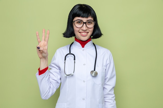 Jovem bonita sorridente com óculos ópticos menina caucasiana com uniforme de médico com estetoscópio gesticulando três com os dedos