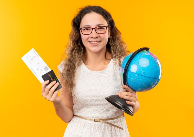 Jovem bonita sorridente de óculos segurando uma passagem de avião, cartão de crédito e globo isolado na parede amarela