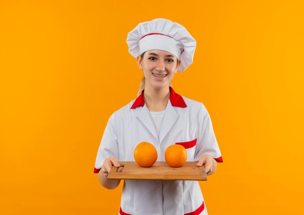 Jovem bonita sorridente em uniforme de chef com aparelho dentário segurando uma tábua de cortar com laranjas isoladas em um espaço laranja