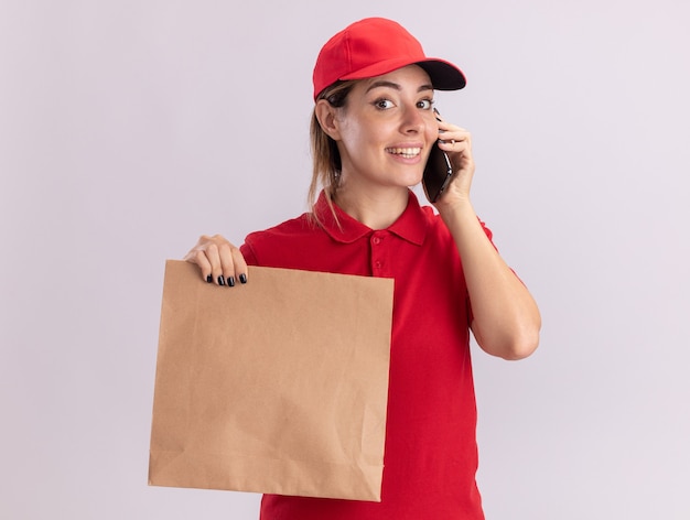 Foto grátis jovem bonita sorridente, entregadora de uniforme, segurando um pacote de papel e falando no telefone, isolado na parede branca