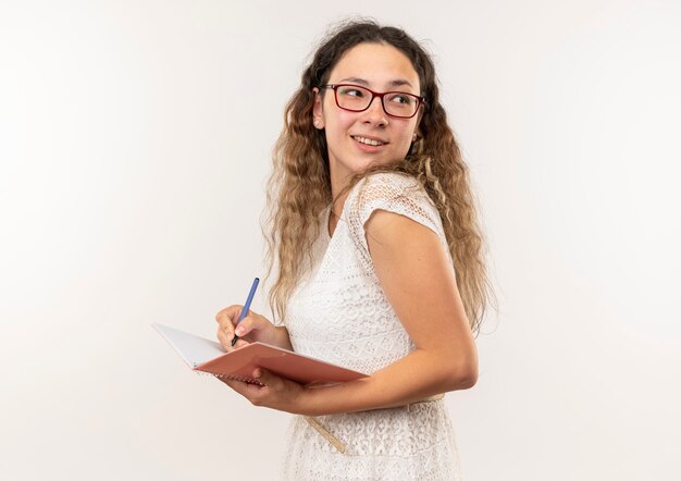 Jovem bonita sorridente, usando óculos e bolsa traseira, em vista de perfil, olhando para trás, escrevendo com uma caneta no bloco de notas isolado na parede branca