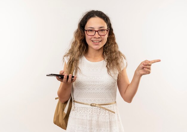 Jovem bonita sorridente usando óculos e bolsa traseira segurando o celular apontando para o lado isolado na parede