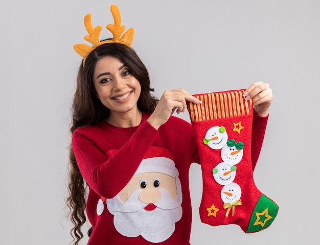 Jovem bonita sorridente usando uma faixa de chifres de rena e um suéter de papai noel segurando uma meia de natal isolada na parede branca