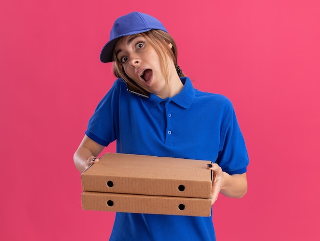 Jovem bonita surpresa, entregadora de uniforme, segurando caixas de pizza e falando no telefone rosa