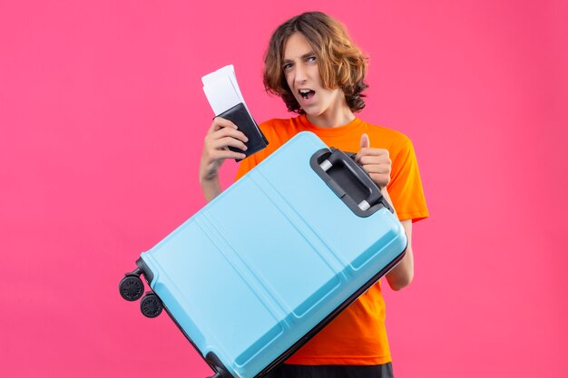 Jovem bonitão de camiseta laranja segurando a mala de viagem e passagens aéreas sorrindo alegremente positivo e feliz em pé