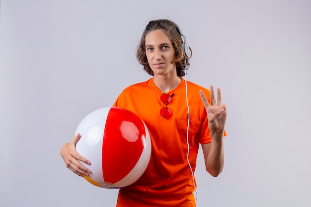 Jovem bonitão de camiseta laranja, segurando uma bola inflável com fones de ouvido, mostrando o número três, sorrindo em pé