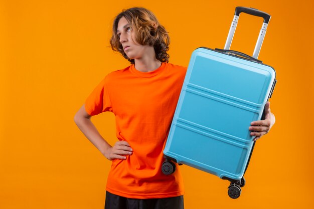 Jovem bonitão na camiseta laranja segurando a mala de viagem, olhando de lado entediado e cansado em pé