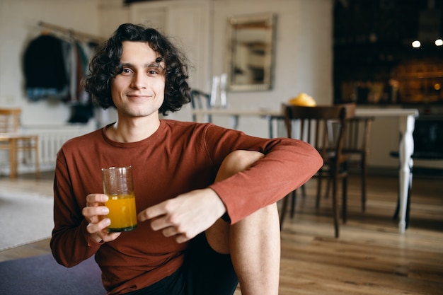 Jovem bonito alegre sentado no chão, comendo laranja fresca no café da manhã, tomando suco em jejum, sorrindo feliz para a câmera