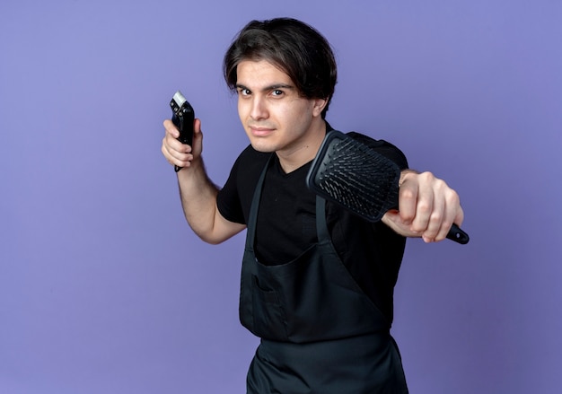 Jovem bonito barbeiro confiante de uniforme segurando o pente com uma tesoura de cabelo isolada no fundo azul