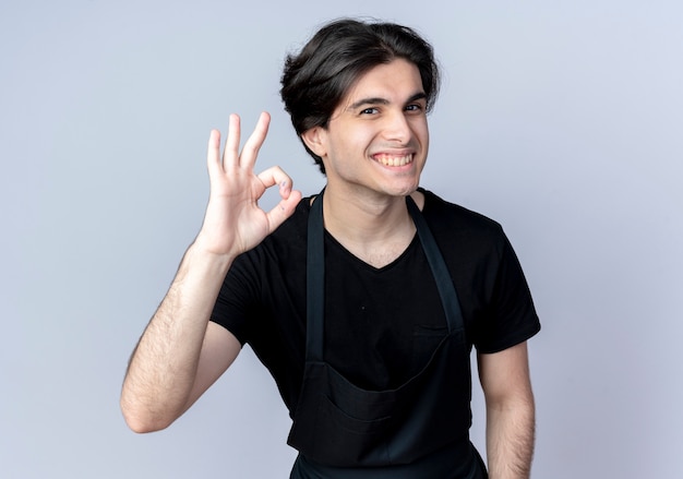 Jovem bonito barbeiro de uniforme sorridente, mostrando o gesto certo isolado no branco