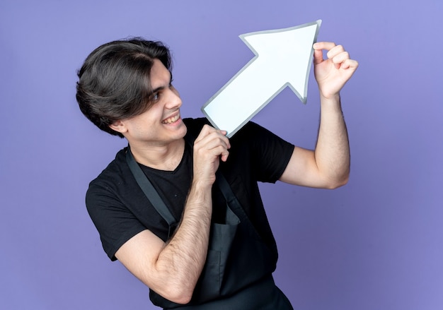 Foto grátis jovem bonito barbeiro de uniforme sorridente segurando e olhando para a marca de direção do papel isolada no azul