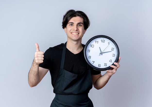 Jovem bonito barbeiro de uniforme sorridente segurando um relógio de parede com o polegar isolado no branco