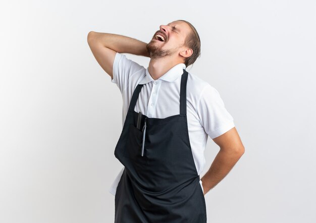 Jovem bonito barbeiro dolorido vestindo uniforme segurando seu pescoço e costas com os olhos fechados, sofrendo de dor isolada no branco com espaço de cópia