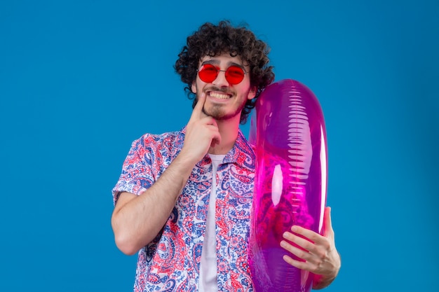 Foto grátis jovem bonito cacheado sorridente usando óculos escuros segurando um anel de natação com o dedo no queixo na parede azul isolada com espaço de cópia