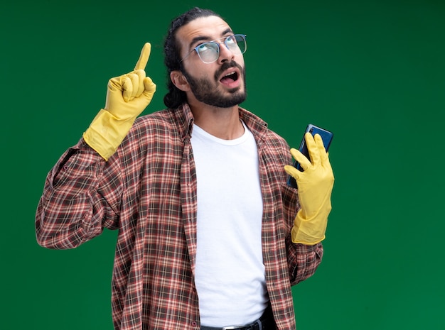 Jovem, bonito, cara de limpeza impressionado, vestindo camiseta e luvas, segurando as pontas do telefone isoladas na parede verde