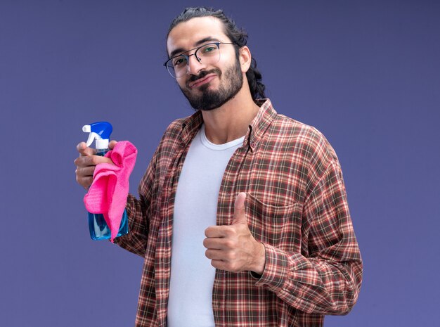 Jovem, bonito, cara de limpeza satisfeito, vestindo uma camiseta, segurando o frasco de spray com um pano aparecendo o polegar isolado na parede azul