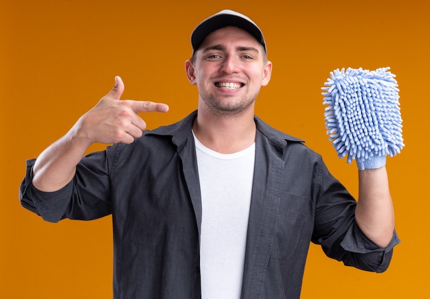 Jovem, bonito, cara de limpeza sorridente com camiseta e boné segurando e apontando para um pano de limpeza isolado na parede laranja