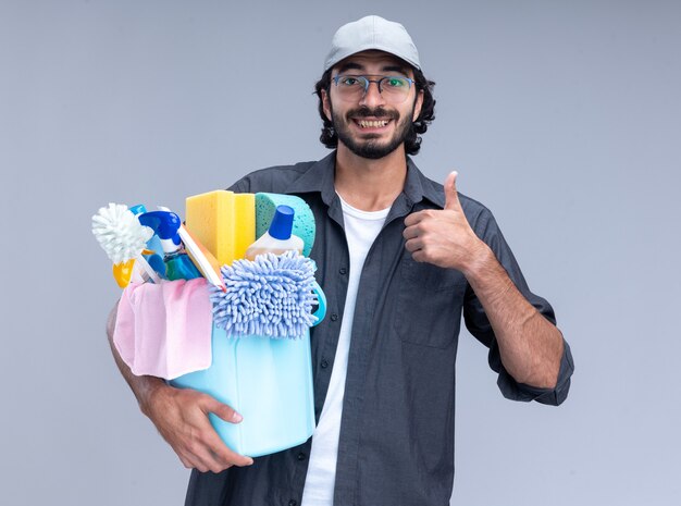 Jovem, bonito, cara de limpeza sorridente, vestindo camiseta e boné, segurando um balde de ferramentas de limpeza aparecendo o polegar isolado na parede branca