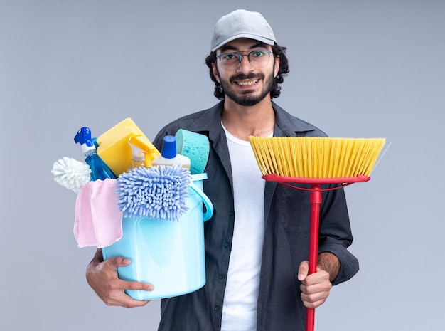 Jovem, bonito, cara de limpeza sorridente, vestindo camiseta e boné, segurando um balde de ferramentas de limpeza com o esfregão isolado na parede branca