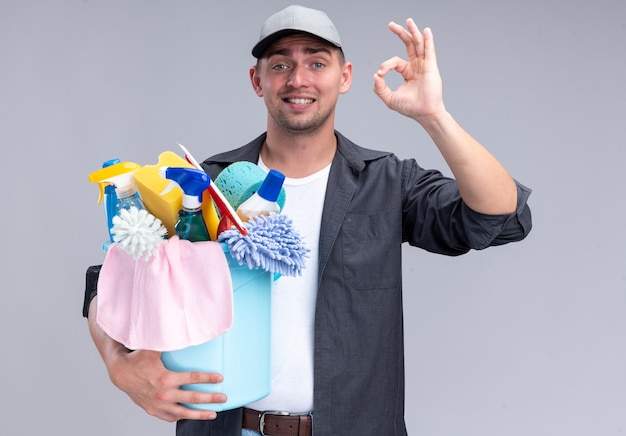 Jovem, bonito, cara de limpeza sorridente, vestindo camiseta e boné, segurando um balde de ferramentas de limpeza, mostrando um gesto de ok isolado na parede branca