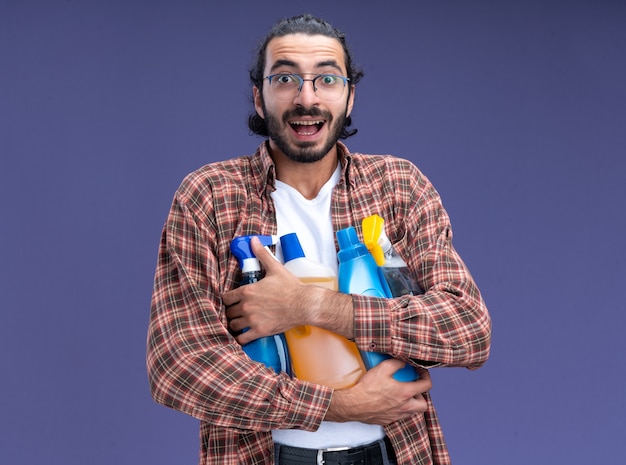 Jovem, bonito, cara de limpeza sorridente, vestindo uma camiseta segurando ferramentas de limpeza isoladas na parede azul